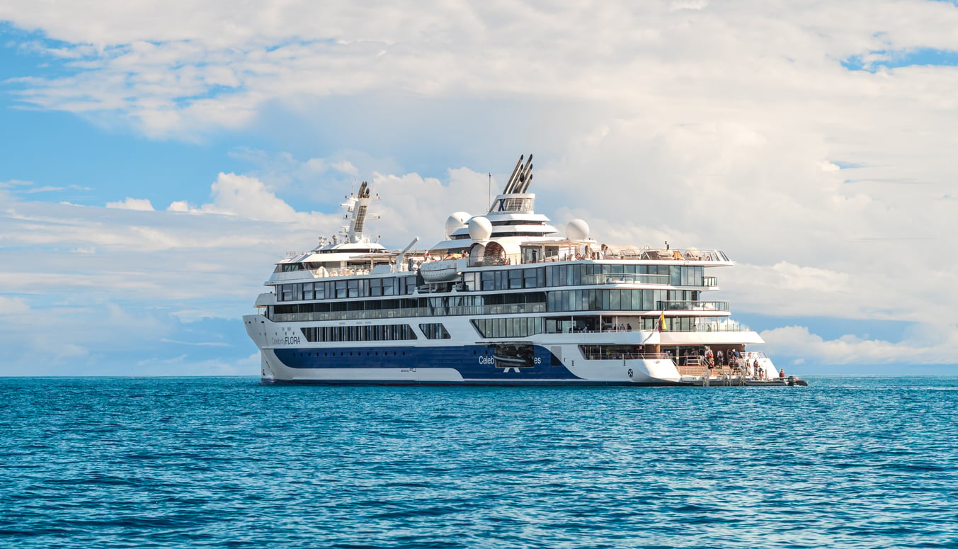 A modern white and blue luxury cruise ship is anchored on calm blue ocean water under a partly cloudy sky.