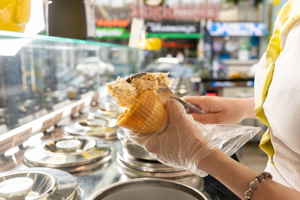 A person wearing a plastic glove scoops ice cream into a waffle cone at an ice cream shop, with metal tubs of various flavors in the display case—tempting customers despite the notoriously overpriced products.