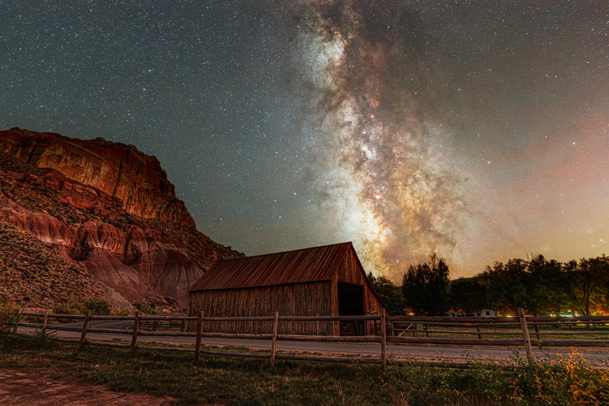 A rustic wooden barn sits beneath a starry night sky with the Milky Way clearly visible, surrounded by a wooden fence, rocky hills, and sparse trees.