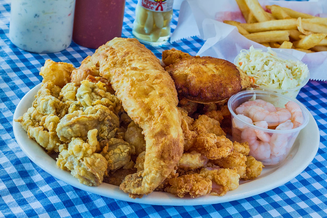 A plate of fried seafood including fish, shrimp, oysters, and a crab cake, served with coleslaw and fries on a blue checkered tablecloth. Condiments and a drink are visible in the background.