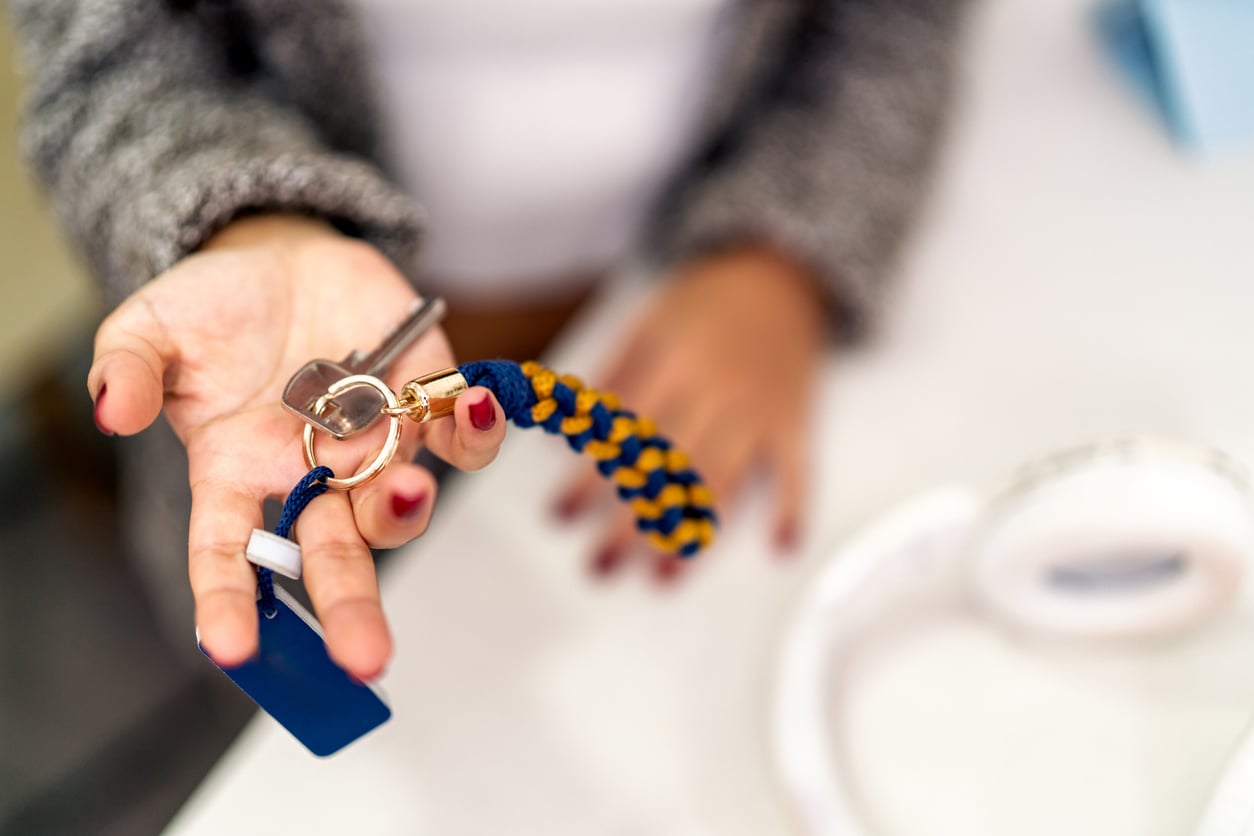 A person with red-painted nails holds out a set of keys with a blue and yellow braided keychain and a blue key tag, against a blurred indoor background.