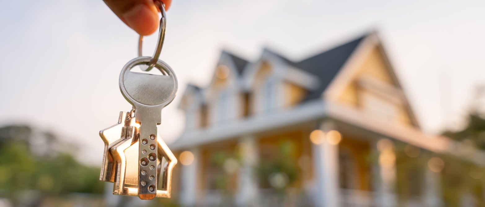 A hand holding a keychain with house-shaped keys in the foreground, with a blurred two-story house in the background, suggesting a new home or real estate concept.