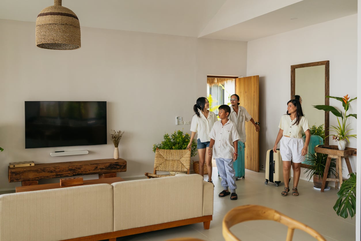 A family of four enters a modern, bright living room with neutral decor, plants, and a flat-screen TV. One person holds a suitcase as they all look around, happy and excited to be staying at an Airbnb.