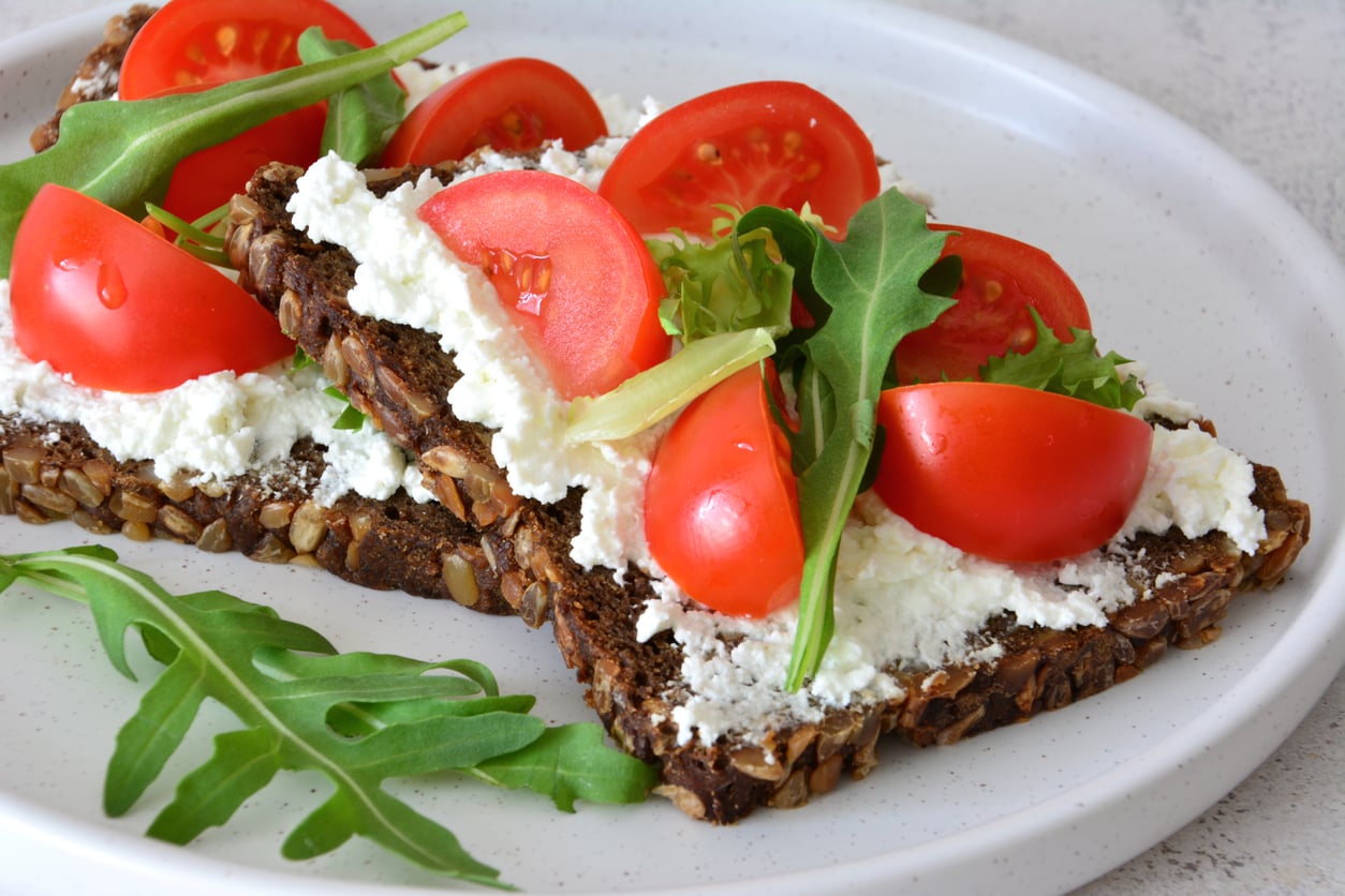Two slices of seeded whole grain bread topped with ricotta cheese, cherry tomato halves, and fresh arugula—an easy, delicious option for those seeking fresh ground beef alternatives, served on a white plate with extra arugula on the side.