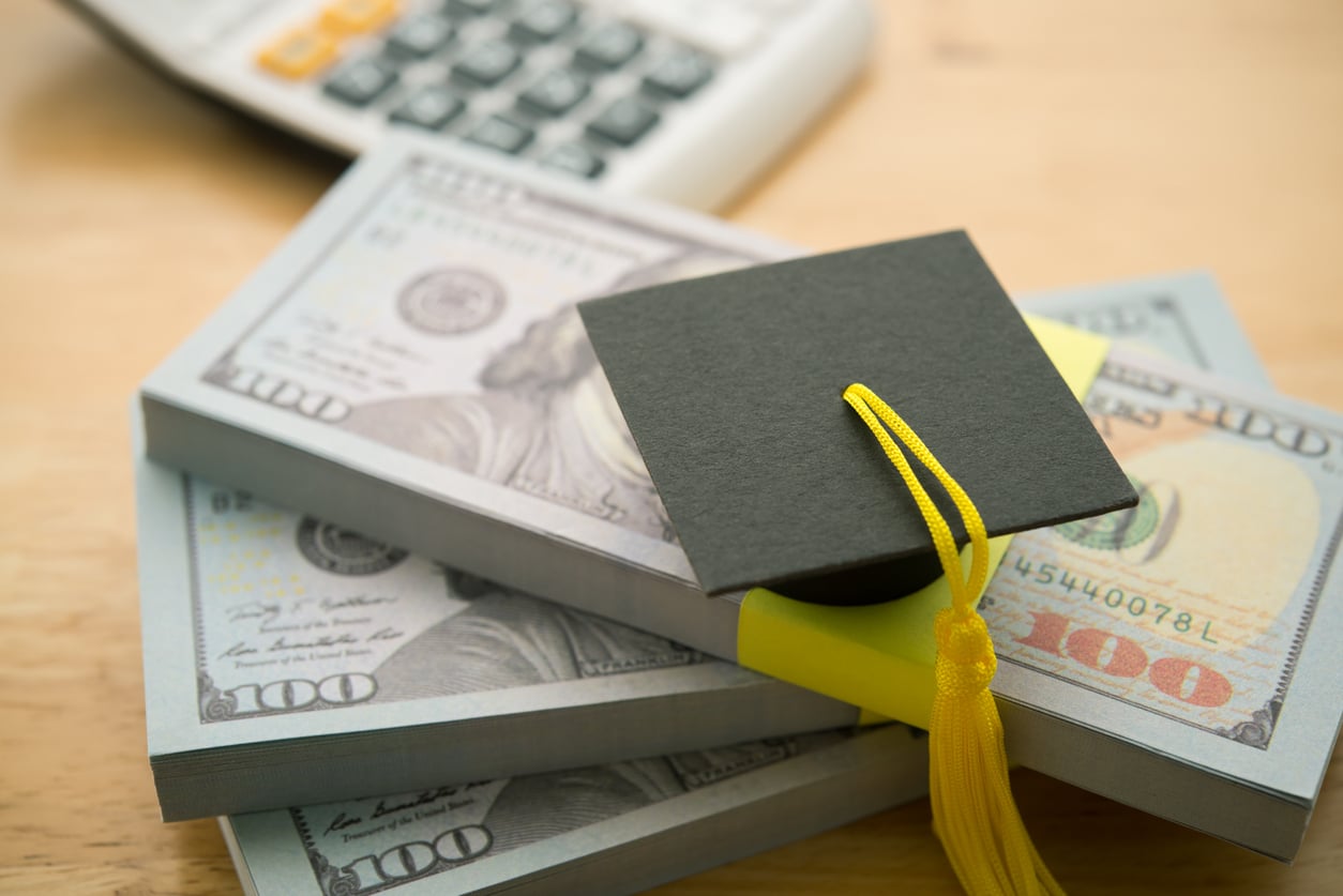 A small graduation cap sits on top of stacks of 100-dollar bills, with a calculator blurred in the background, symbolizing student loans or education costs.