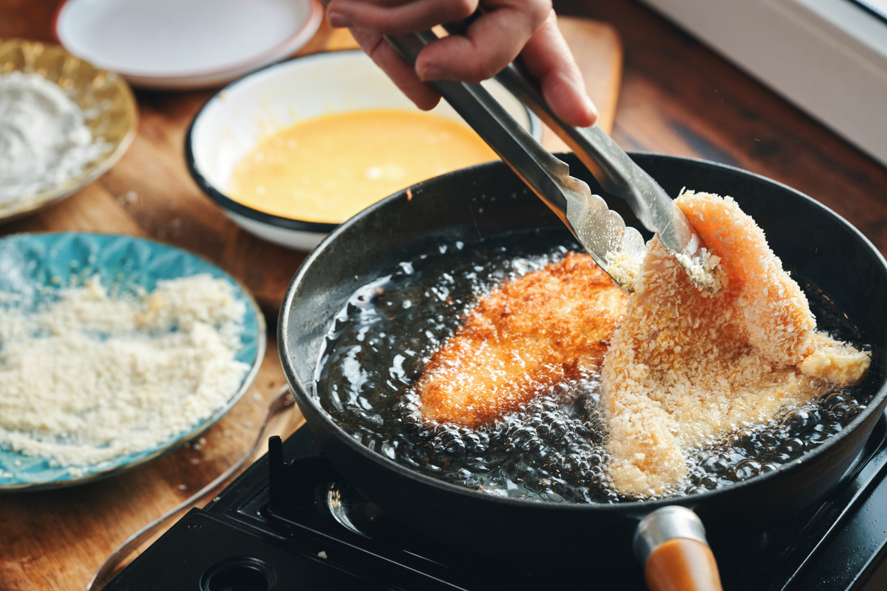 A hand uses tongs to fry breaded cutlets in a pan of hot oil, with bowls of flour, beaten eggs, and breadcrumbs on the side for coating.