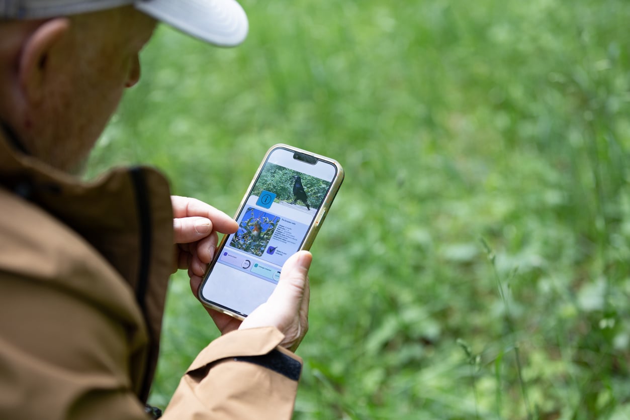 A person in an outdoor setting enjoys budget-friendly hobbies, using a smartphone with a map or navigation app open, pointing at the screen while sitting or kneeling on green grass.