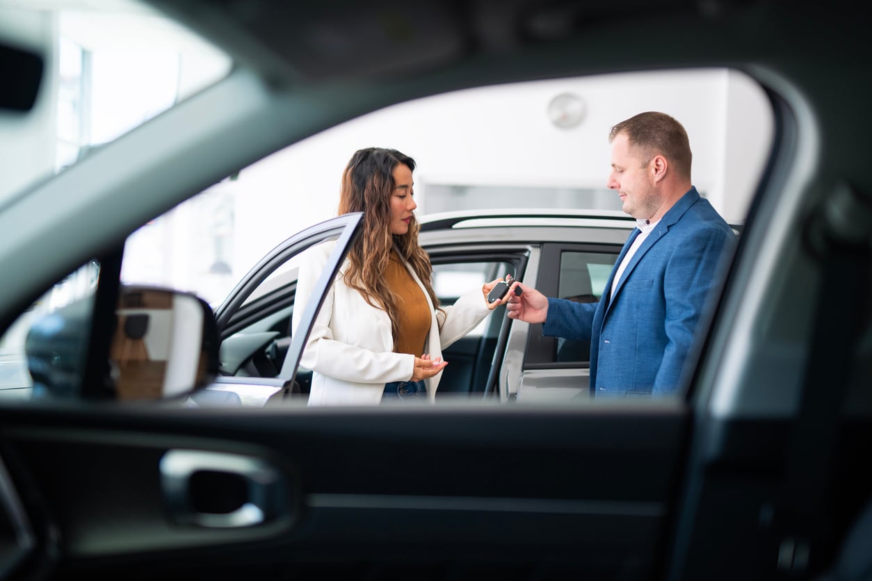 A woman receives car keys from a man in a blue blazer at a car dealership, standing next to an open car door, viewed through the window of another car.