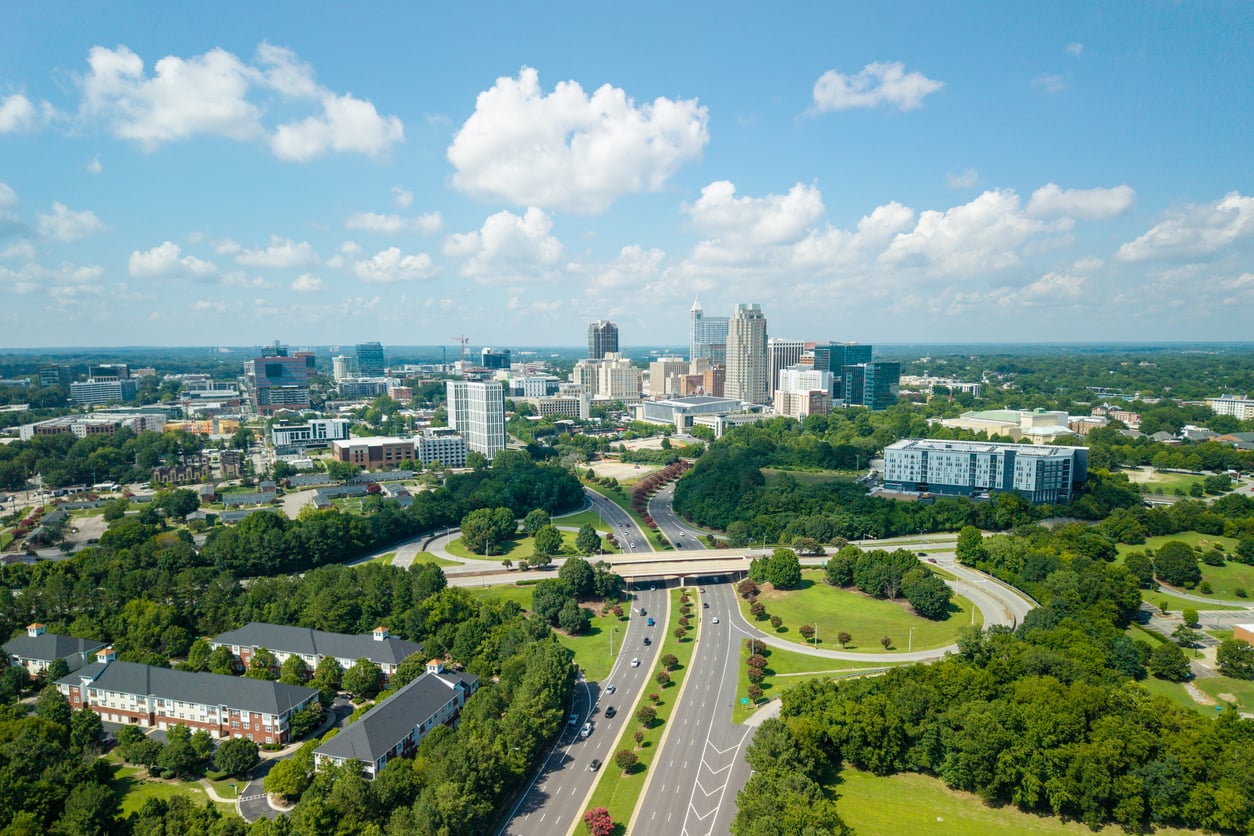 Aerial view of a city skyline in one of the top Small Business States, with tall buildings in the background, a highway with several cars running through green trees and parks, and scattered clouds in a blue sky.