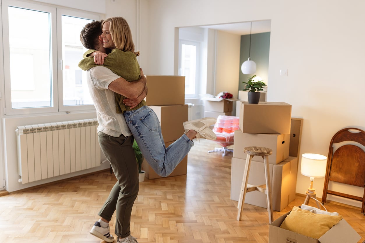 A happy couple hugs and smiles in a bright room filled with moving boxes, suggesting they are moving into a new home. One person lifts the other off the floor as they celebrate together.