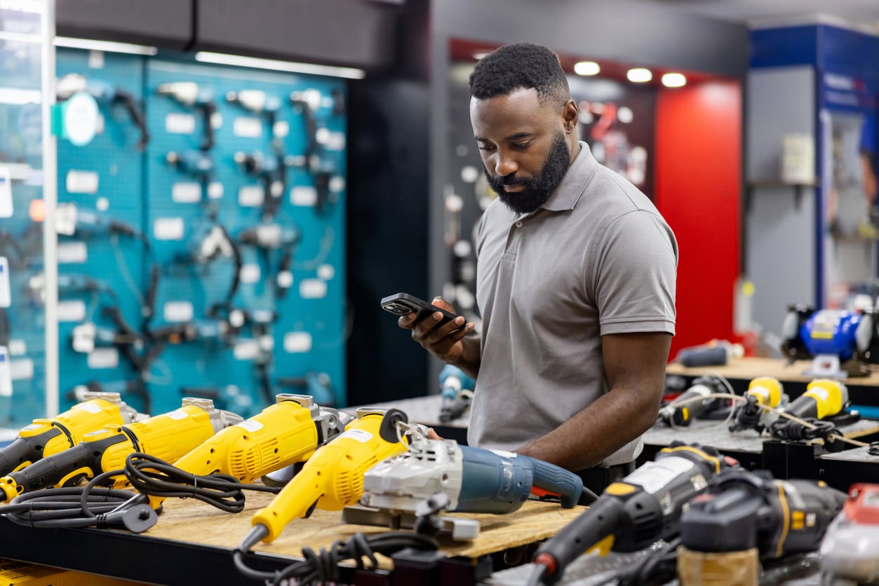A man in a gray polo shirt stands at a hardware store counter, eyeing power tools while holding his phone—perhaps considering which products men skip buying among the yellow and black electric tools displayed before him.
