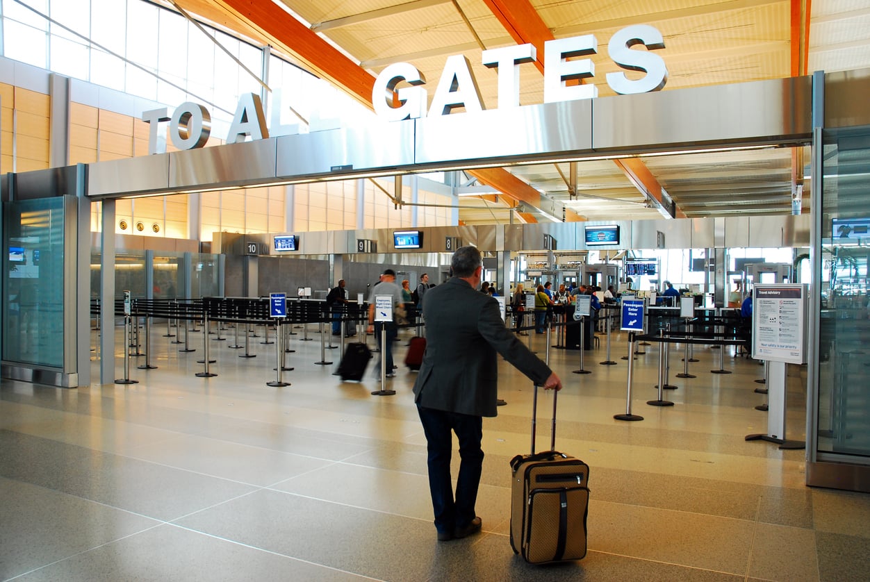A man in a suit pulls a suitcase toward the security checkpoint at one of the convenient airports, heading to the area labeled "TO ALL GATES." Other travelers and security lines are visible ahead.