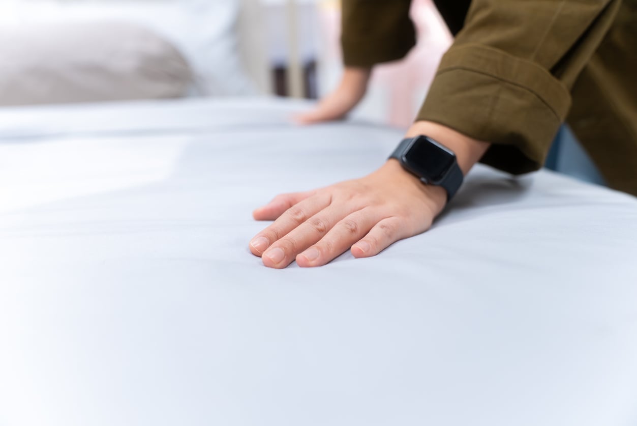 A person wearing a smartwatch and an olive green sleeve presses their hand on a smooth, white bed sheet, testing its softness—one of those products men still buy for comfort. The background is softly blurred.