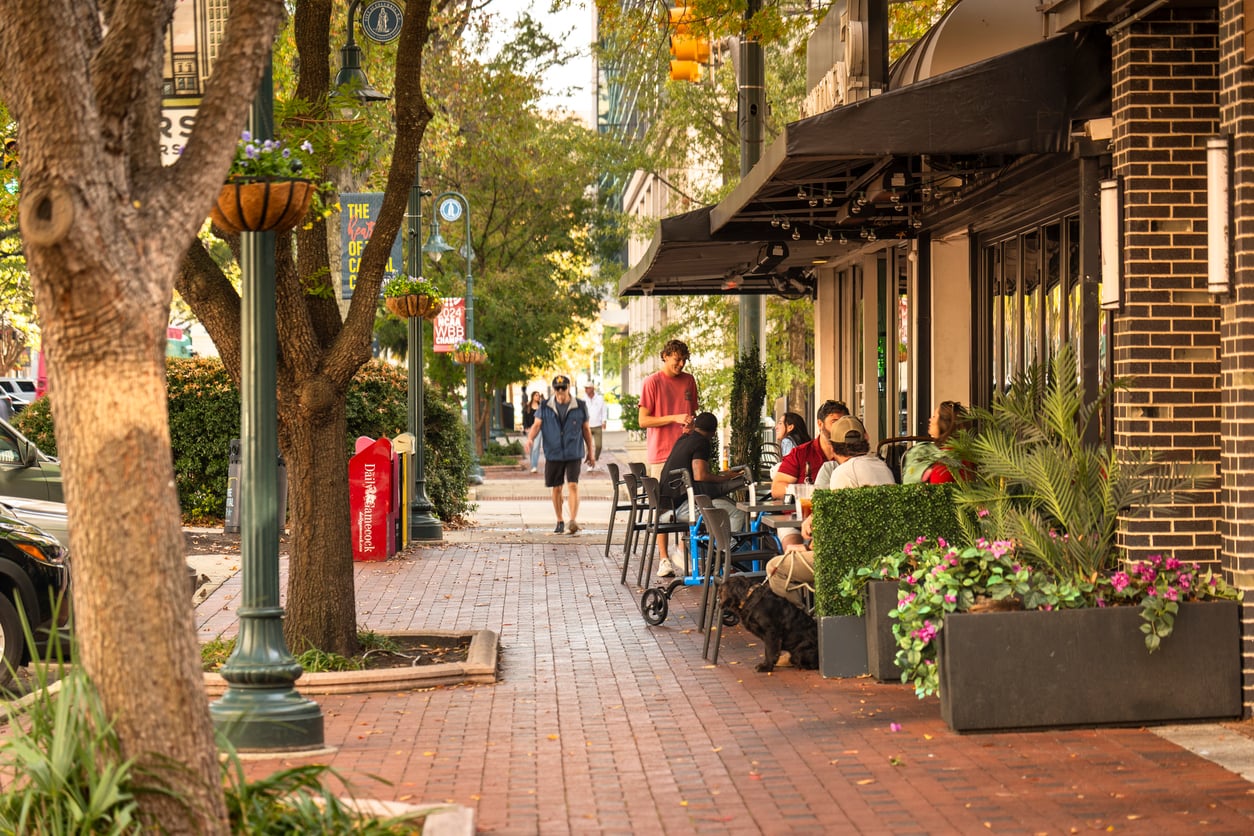 People dine at outdoor tables along a tree-lined brick sidewalk in one of the vibrant Small Business States, while others walk in the background. Potted plants and street lamps decorate the lively urban scene.