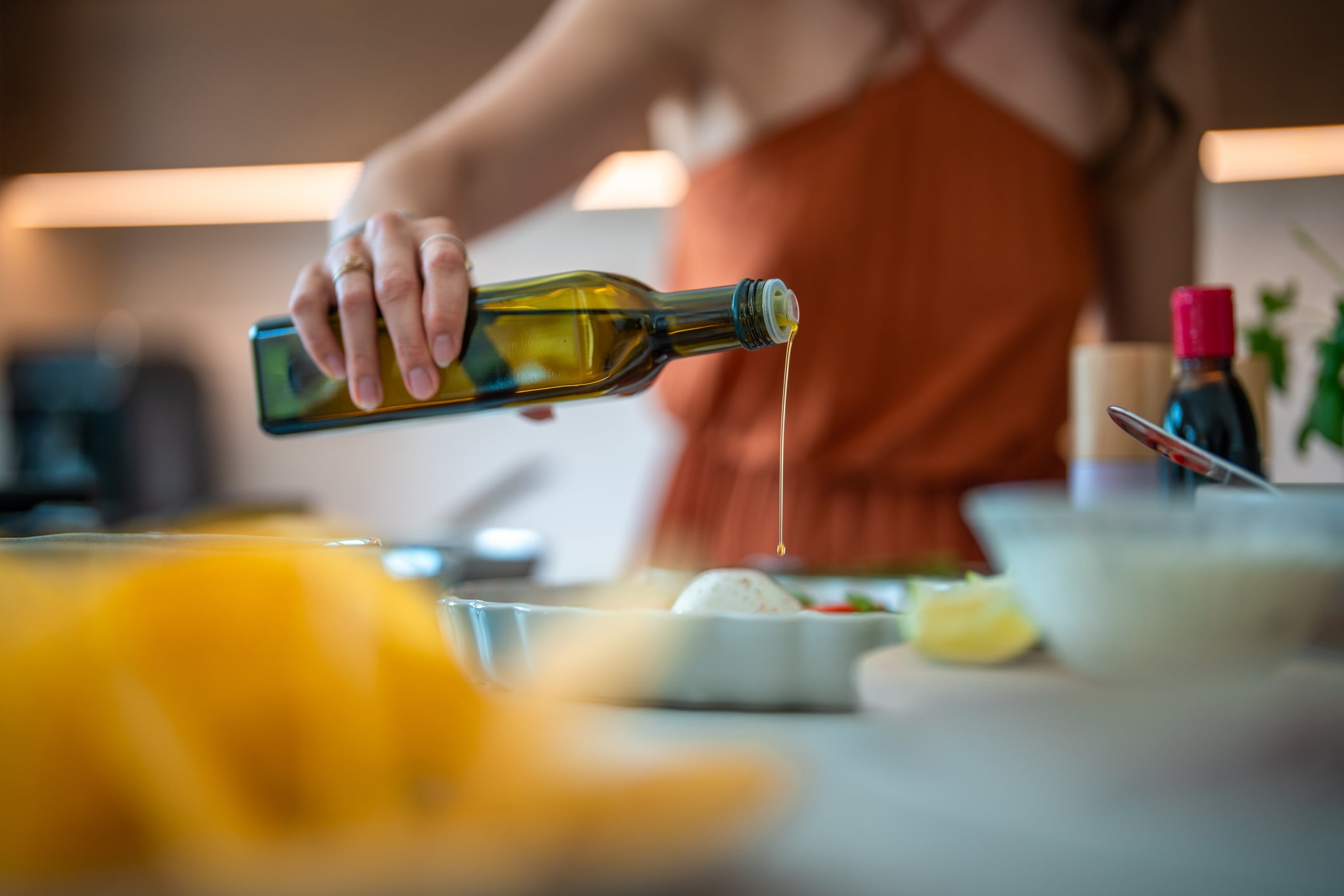 A woman in a sleeveless brown dress pours olive oil from a bottle onto food in a kitchen, with dishes and ingredients like lemon and sauces visible on the counter.