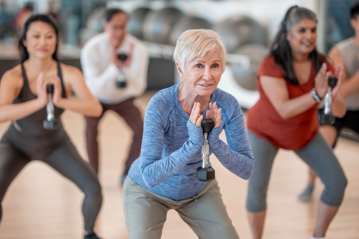 A group of adults in workout clothes perform squats with dumbbells during a fitness class in a gym. The focus is on an older woman in front, smiling as she exercises—highlighting the connection between physical activity and mental health.