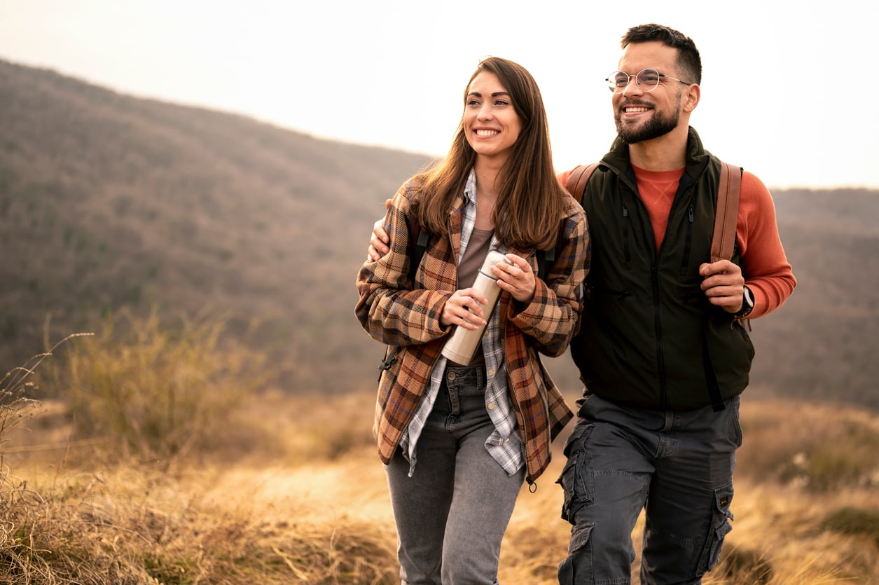 A smiling couple, dressed in casual outdoor clothing, enjoys one of their favorite budget-friendly hobbies as they walk together across a grassy landscape with hills in the background. The woman holds a water bottle while the man has an arm around her shoulder.