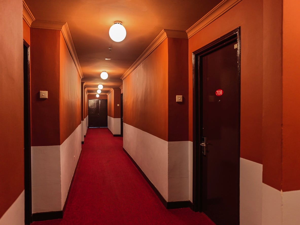 A long, dimly lit hotel hallway with red carpet, orange and white walls, several closed doors, and round ceiling lights. Room 708 is visible on the right side of the corridor. Hotel horror stories.