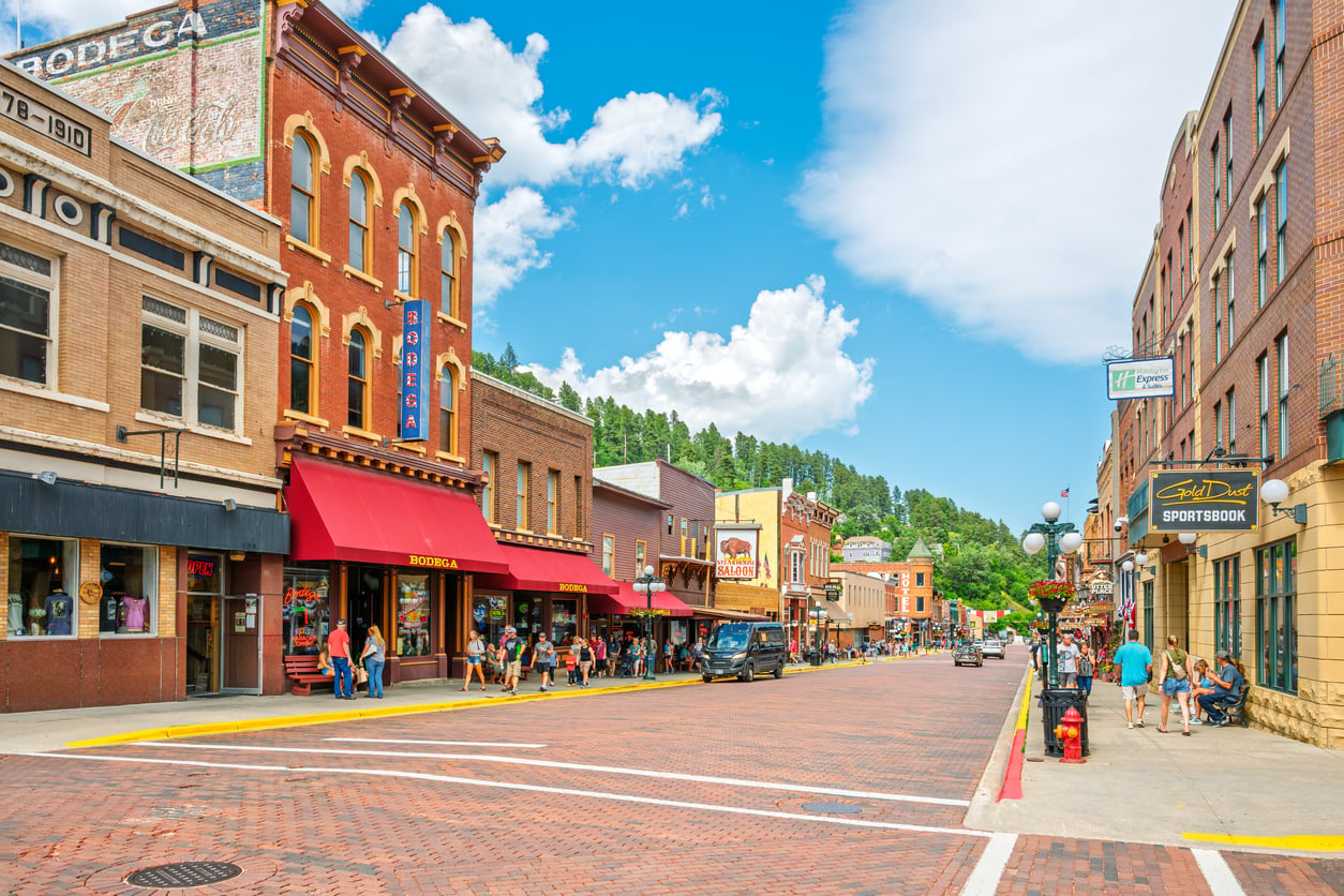 A vibrant downtown street scene with historic brick buildings, shops like Dollar Tree, and restaurants. People walk along sidewalks, and cars are parked by the curb. Green trees and a bright blue sky with clouds complete the lively background.