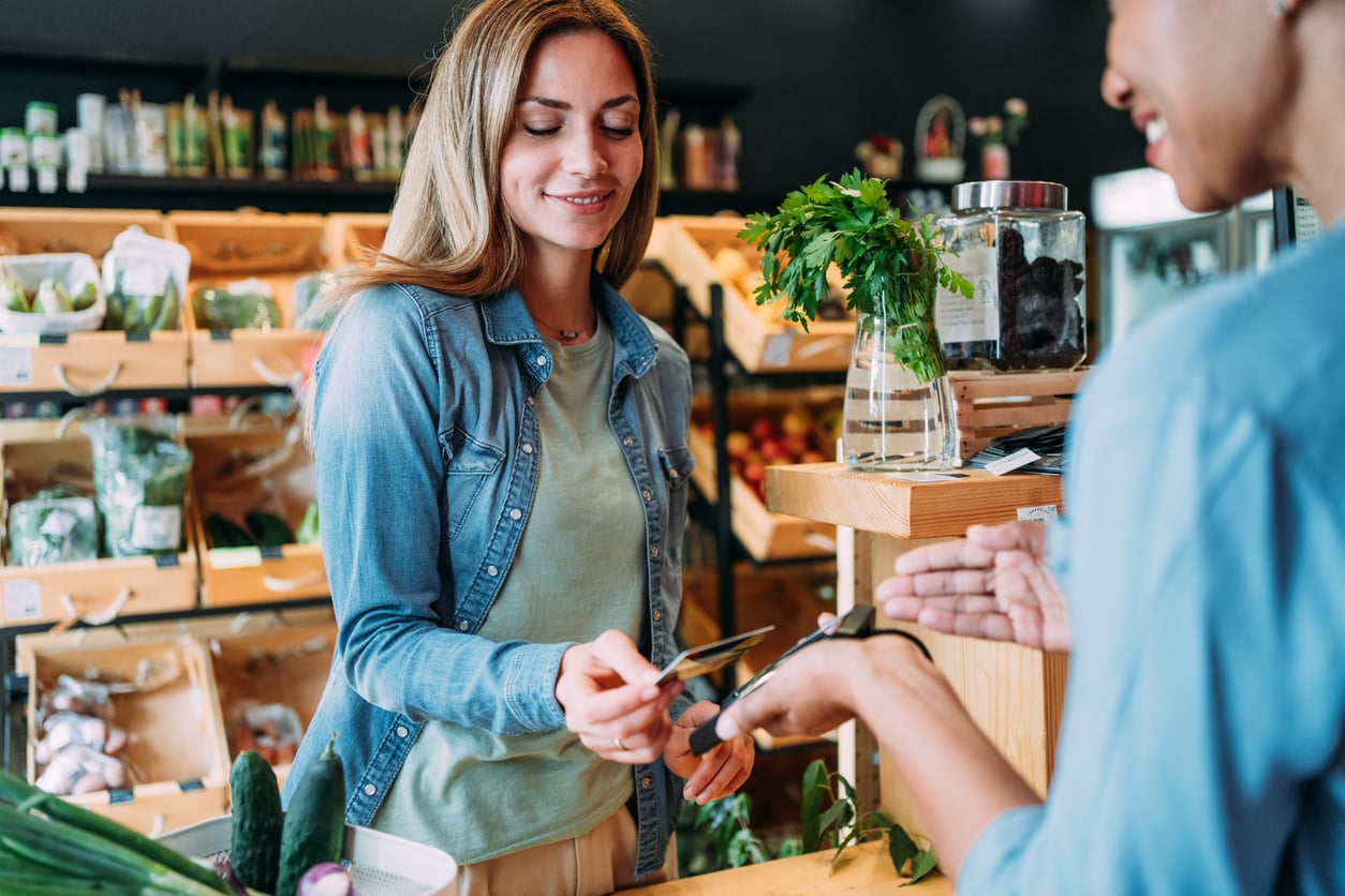 A woman smiles as she pays with a credit card at a grocery store counter, while the cashier holds a payment terminal. Fresh produce and herbs are visible on the counter and shelves in the background.