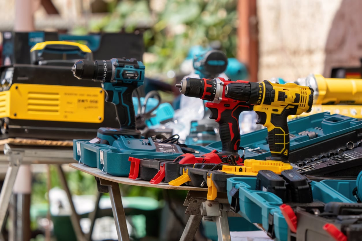 A variety of power drills and toolboxes are displayed on a table outdoors, with other assorted tools and equipment visible in the background.