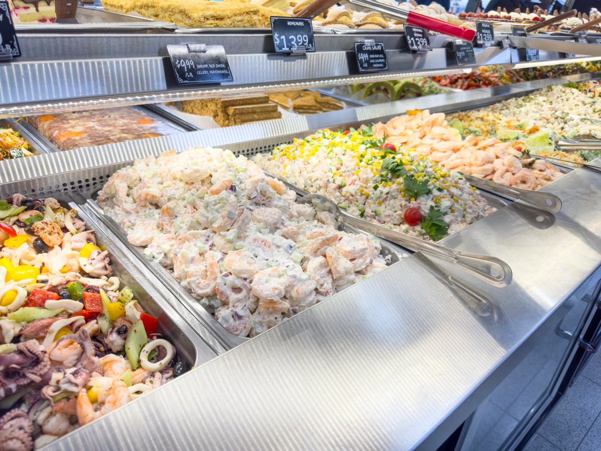A deli counter displays a variety of prepared salads, including seafood and vegetable mixes, arranged in metal trays with serving tongs under bright lighting. Price tags and labels above each item hint at potentially overpriced products.