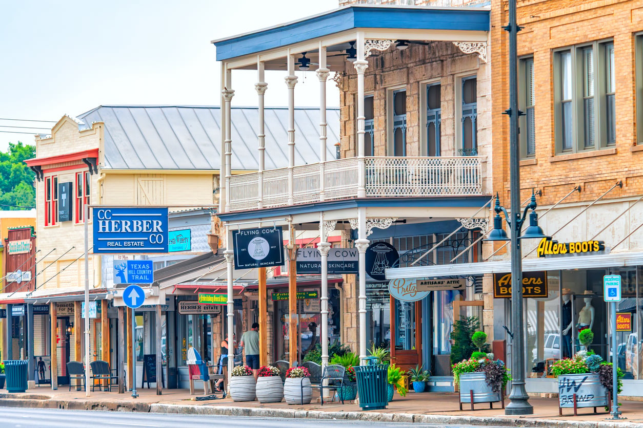 A colorful downtown street scene with historic buildings, various shop signs, outdoor seating, potted plants, and a decorative balcony. Signs include CC Herber Real Estate and Allen’s Boots.