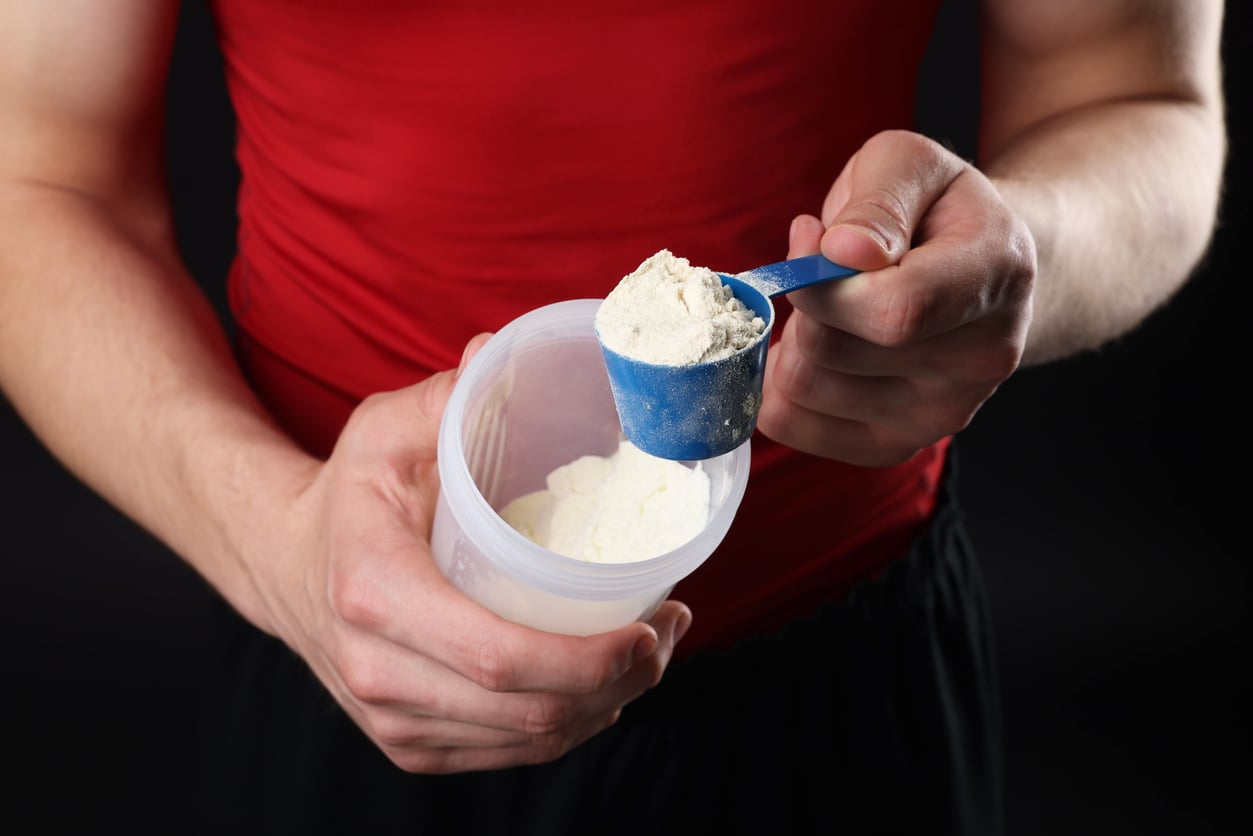 A person in a red shirt scoops protein powder from a container using a blue scoop—one of those classic products men still buy—with both hands visible against a dark background.