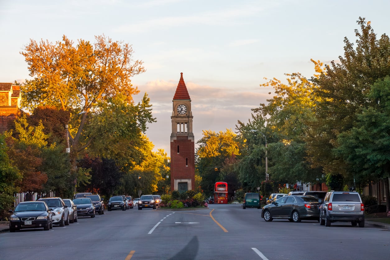 A tree-lined street with parked cars leads to a red-brick clock tower with a pointed roof at sunset. The sky is partly cloudy, and trees have green and golden foliage. Retirement travel destinations. 