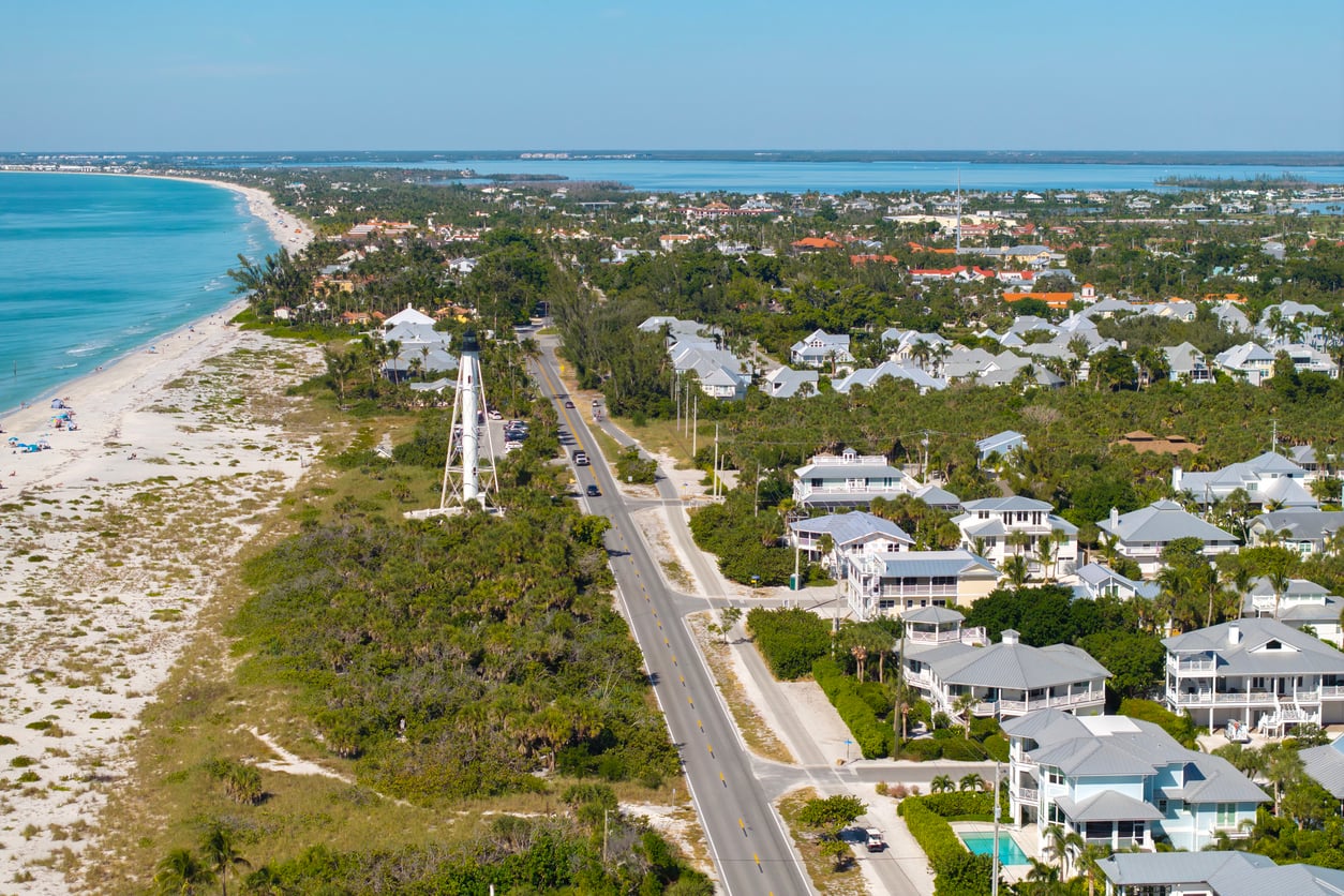 Aerial view of a coastal town with white houses, a lighthouse, and a road running alongside a sandy beach and blue ocean, surrounded by lush greenery under a clear sky.