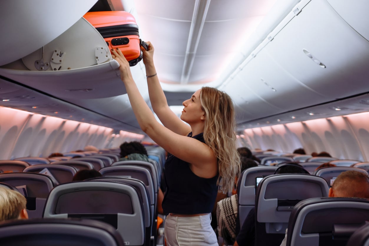 A woman standing in an airplane aisle places an orange suitcase into the overhead compartment while other passengers are seated around her.