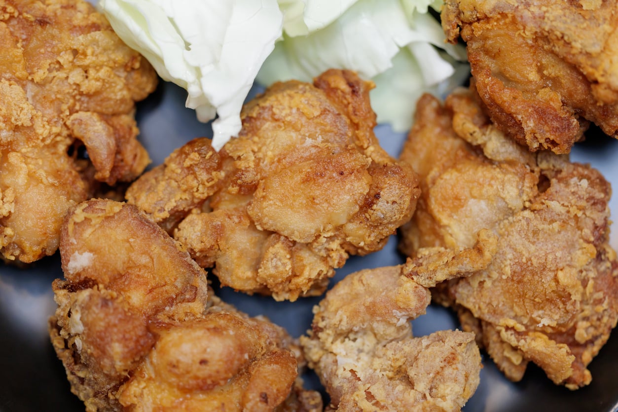 Close-up of several pieces of crispy fried chicken on a dark plate, with a few pieces of fresh, chopped cabbage in the background.