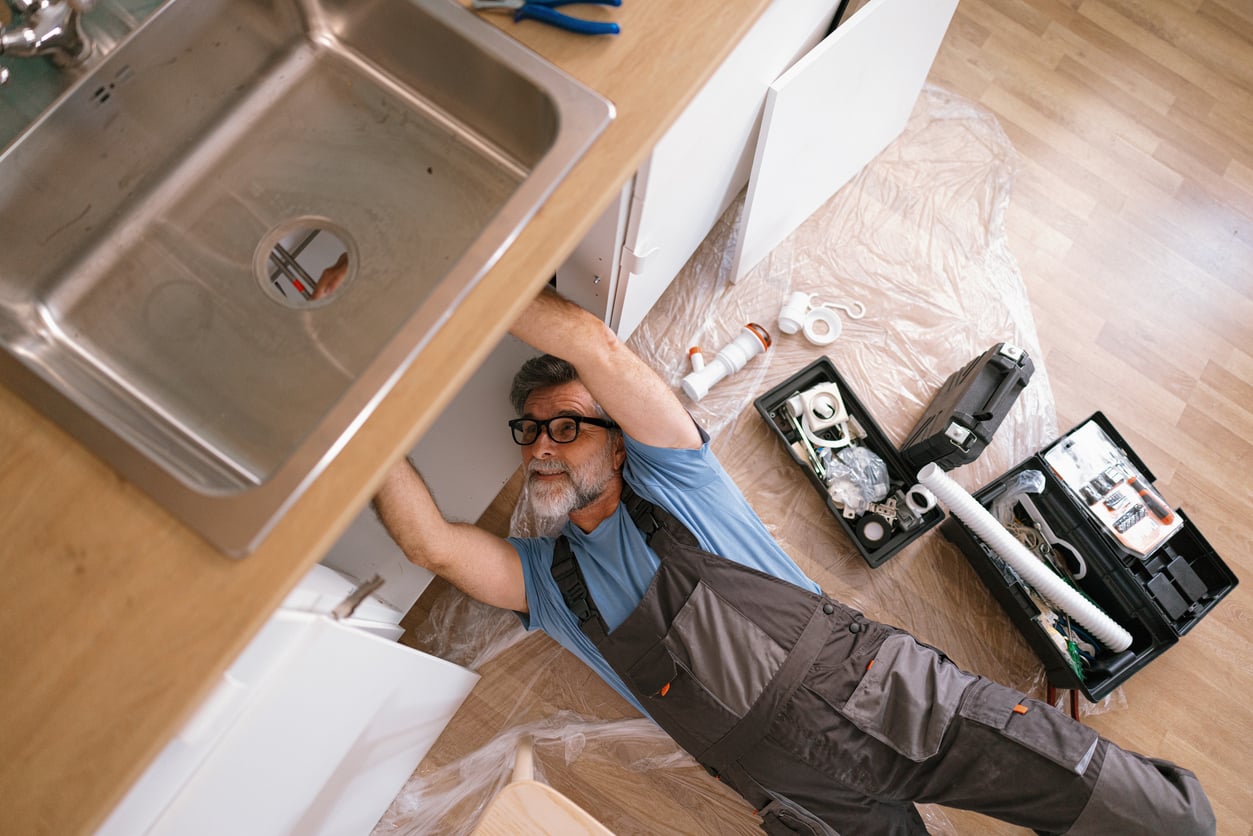 A man in overalls lies on the floor under a kitchen sink, showing off his DIY skills as he works on plumbing repairs. Tools, pipes, and a toolbox are scattered nearby on the plastic-covered floor.
