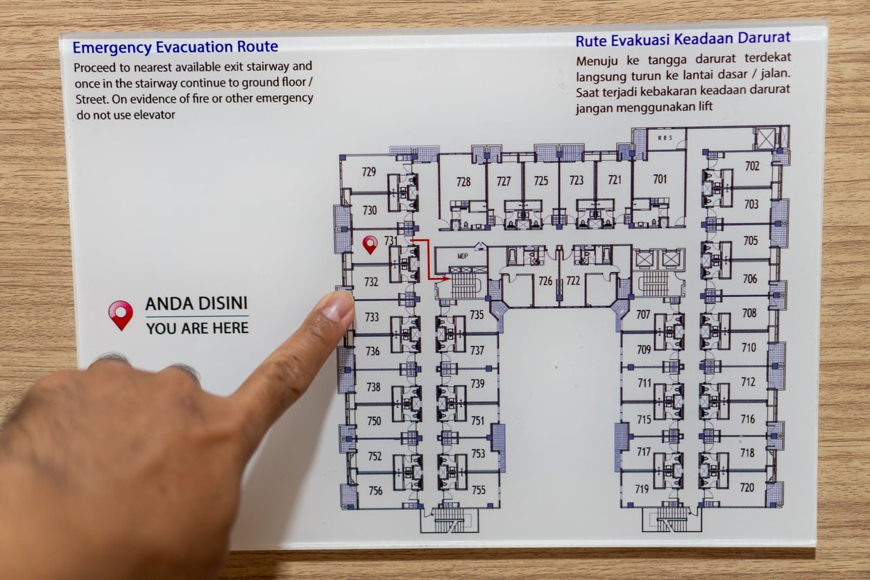 A hand points to a "YOU ARE HERE" marker on a building floor plan, highlighting Hotel Room Safety with emergency evacuation routes in English and Indonesian. Numbered rooms and stairways are clearly marked for safe emergency exits.