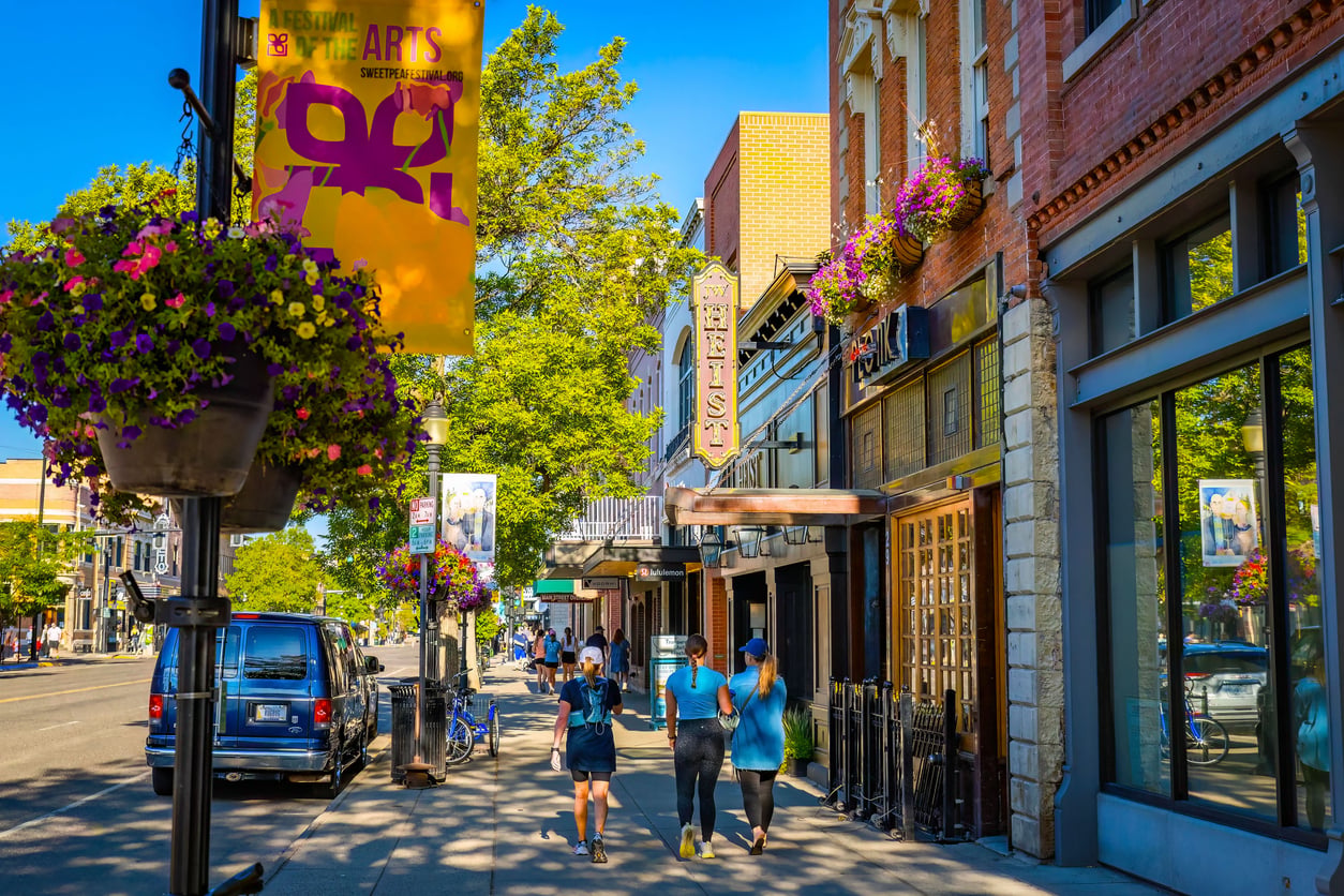 People walk along a lively, sunny street in one of the most affordable college towns, lined with shops, restaurants, and hanging flower baskets. A colorful arts festival banner hangs from a lamppost, with cars parked along the road.