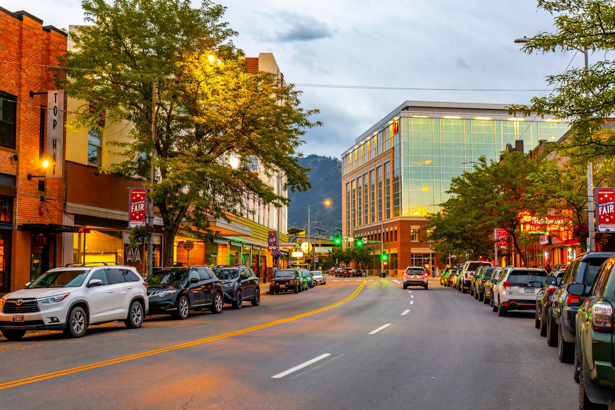 A city street in one of the affordable college towns, lined with parked cars, trees, and buildings. Streetlights and signs glow as dusk falls, with mountains visible in the background beneath a cloudy sky.