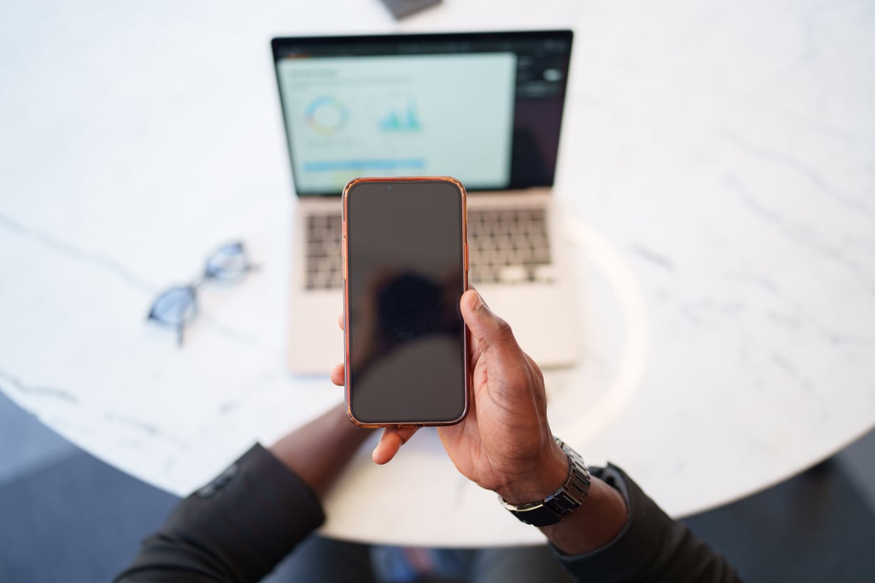 A person holding a smartphone in front of a laptop displaying charts on a white table, with eyeglasses resting nearby—suggesting a moment’s pause to focus on mental health, as the phone screen remains off or blank.
