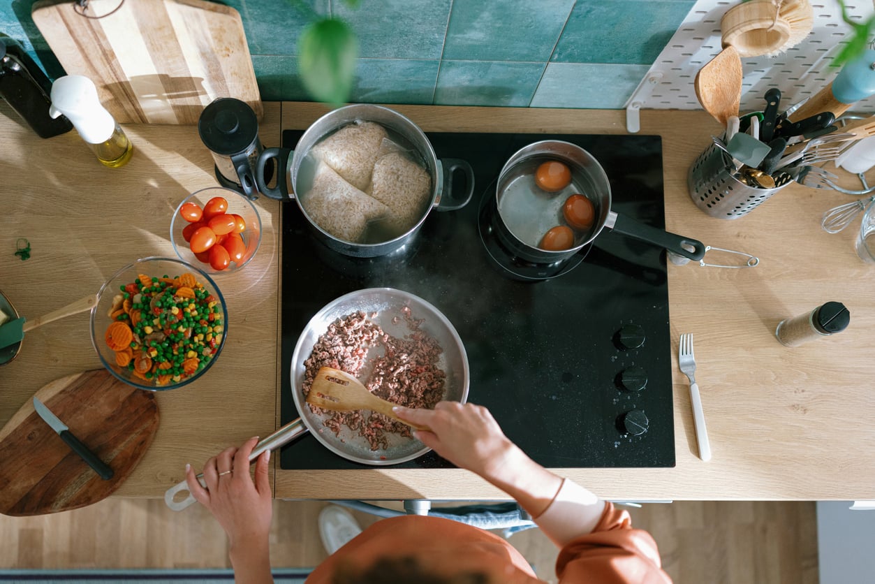 A person cooks ground beef alternatives in a pan on a stovetop, with pots of boiling eggs and rice. Chopped vegetables, fresh tomatoes, and kitchen utensils are arranged on the wooden countertop.