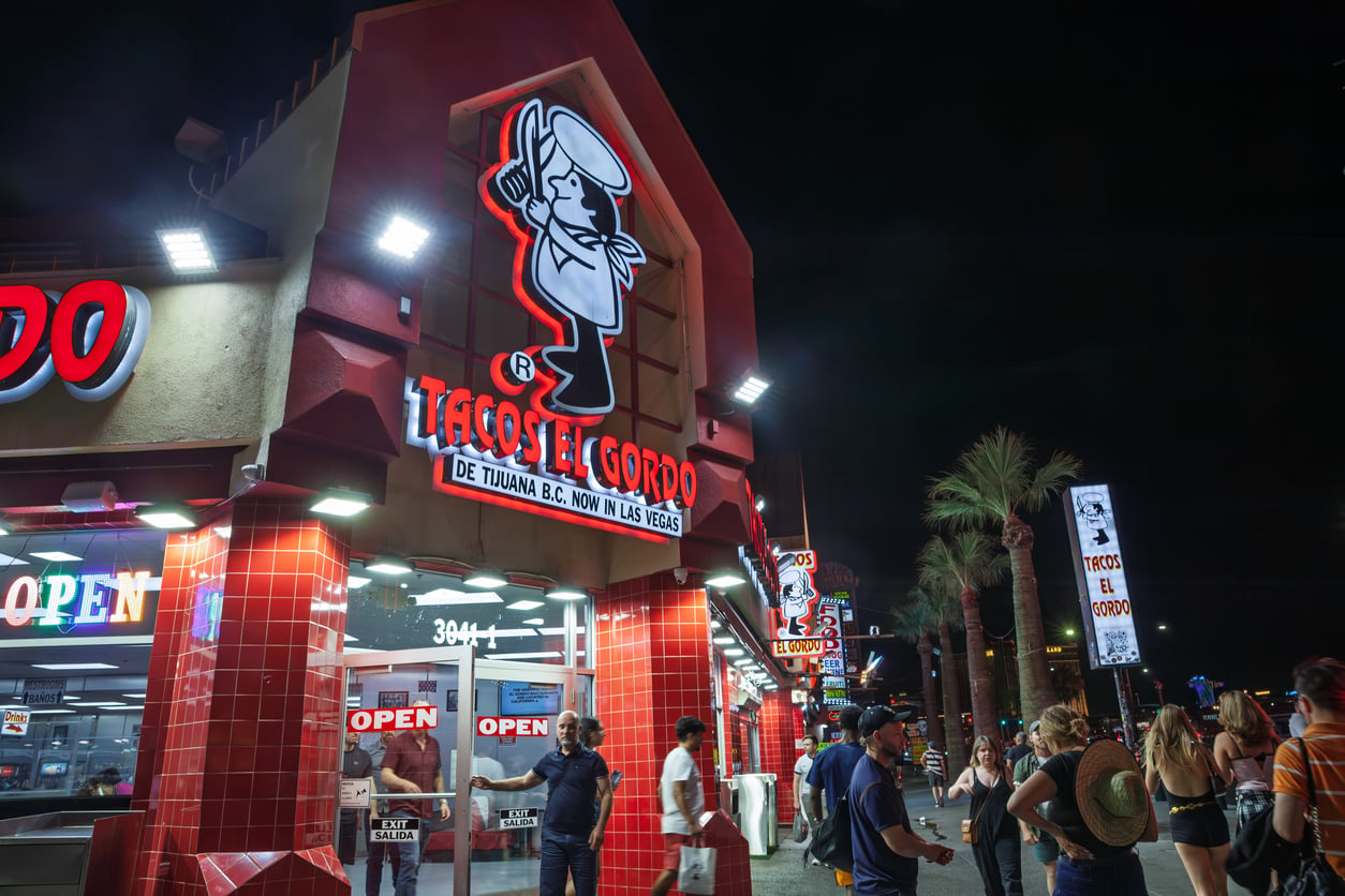 A brightly lit Tacos El Gordo restaurant at night, with people gathered outside. Neon signs, palm trees, and illuminated windows create a lively atmosphere on a busy street—a perfect stop for Las Vegas budget tips seekers.