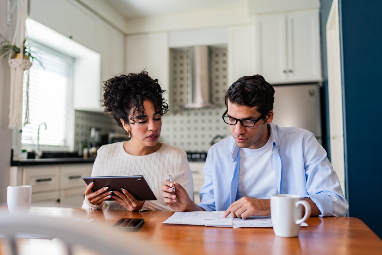 A woman and a man sit at a kitchen table reviewing papers, putting their DIY skills to use. The woman holds a tablet and looks at the man, who is reading documents with a pen. Both appear focused; mugs and a smartphone are on the table.