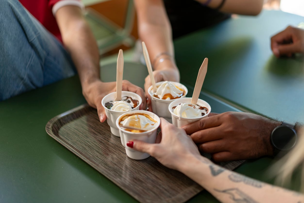 Four people clink their cups of ice cream topped with whipped cream and syrup together over a tray on a green table. Only their hands and arms are visible.