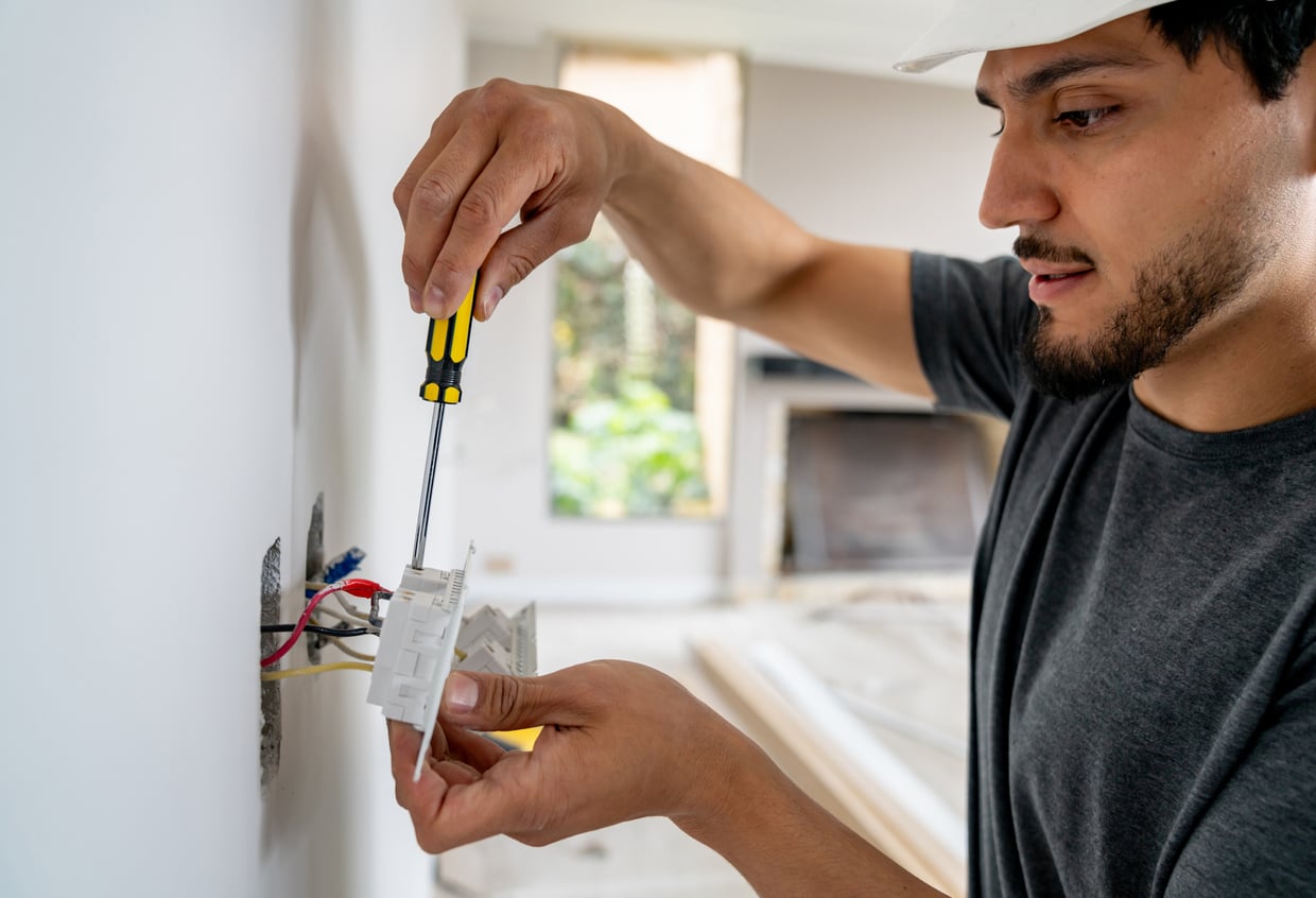 Demonstrating DIY skills, a person wearing a white hard hat uses a screwdriver to install or repair an electrical outlet in a wall, with visible wires and a partially finished room in the background.