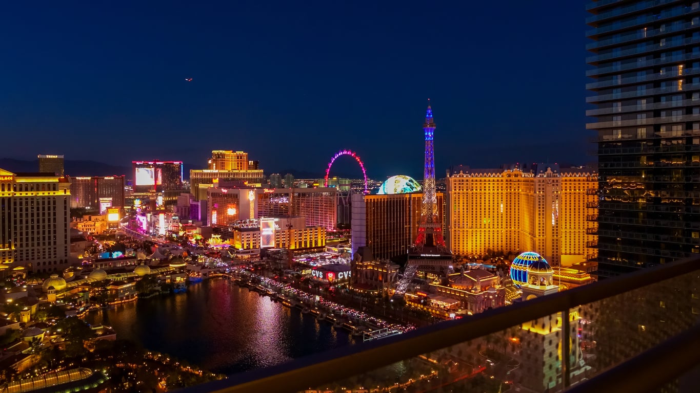 A vibrant nighttime view of the Las Vegas Strip, featuring iconic las vegas attractions like brightly lit hotels, casinos, a replica Eiffel Tower, and the High Roller Ferris wheel, with reflections on nearby water and a deep blue sky.