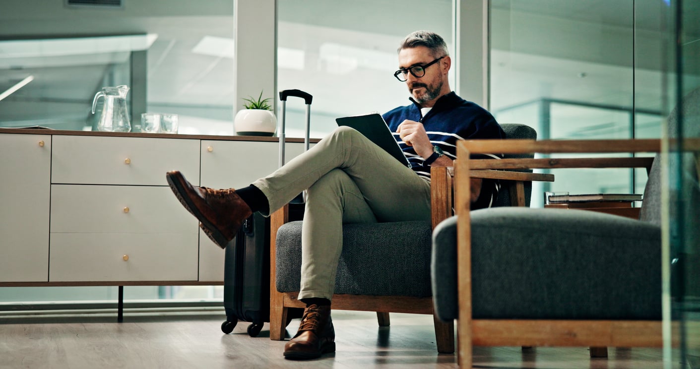 A man with glasses sits in a modern office lounge, reading or writing on a tablet. Dressed in a sweater, pants, and boots, he has a suitcase beside him—perhaps staying at an Airbnb. A cabinet and plant are in the background.