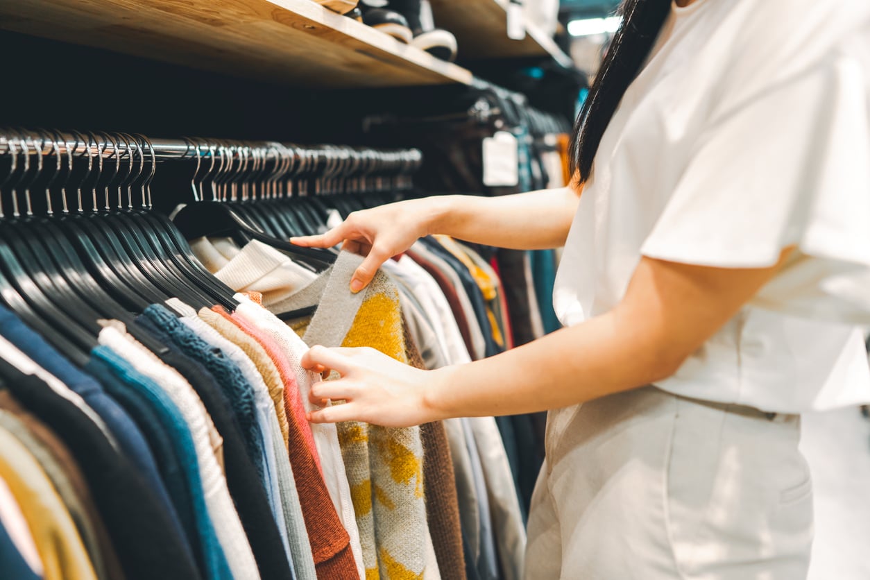 A person browses through a rack of colorful sweaters and shirts in a clothing store, using both hands to separate the hangers and examine the garments.