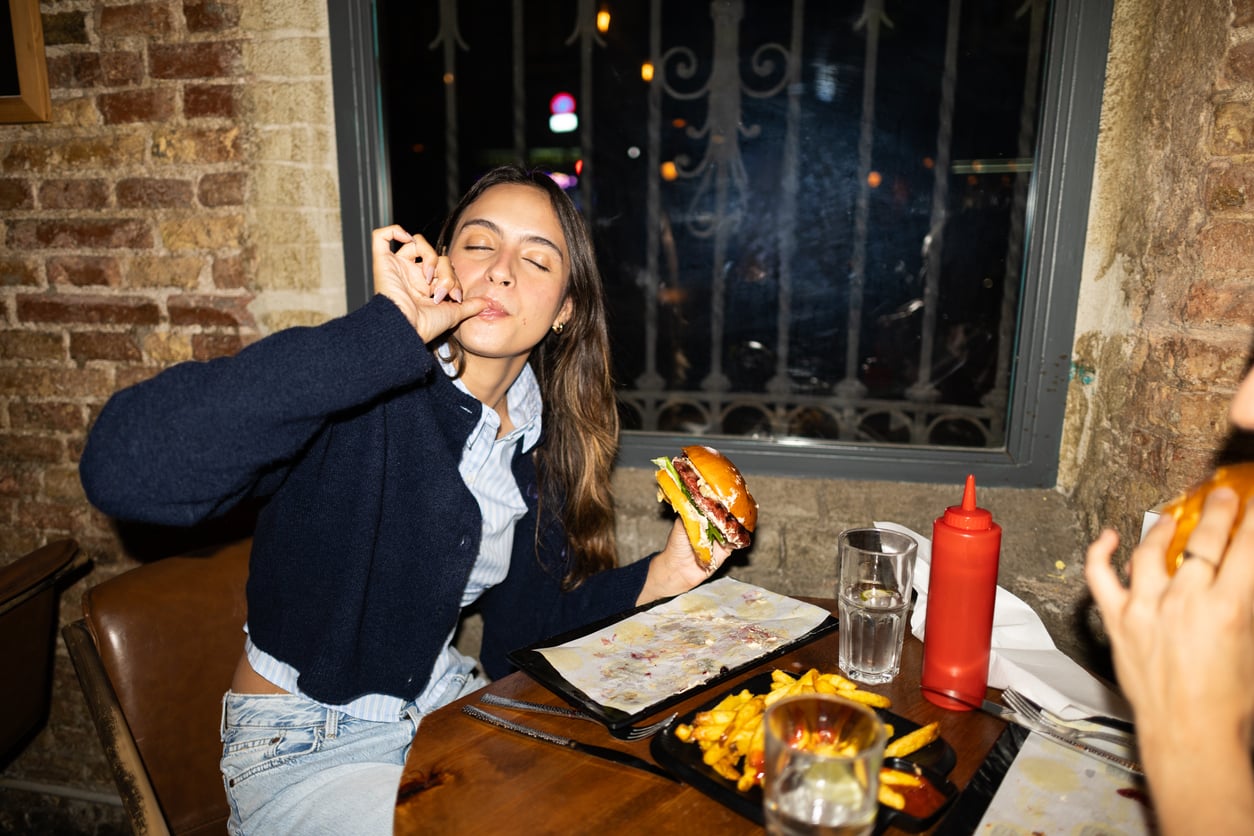A woman sits at a restaurant table, eyes closed, smiling and making an "okay" gesture with her hand while holding a burger. On the table are fries, drinks, and a red ketchup bottle. Another person holds a burger.