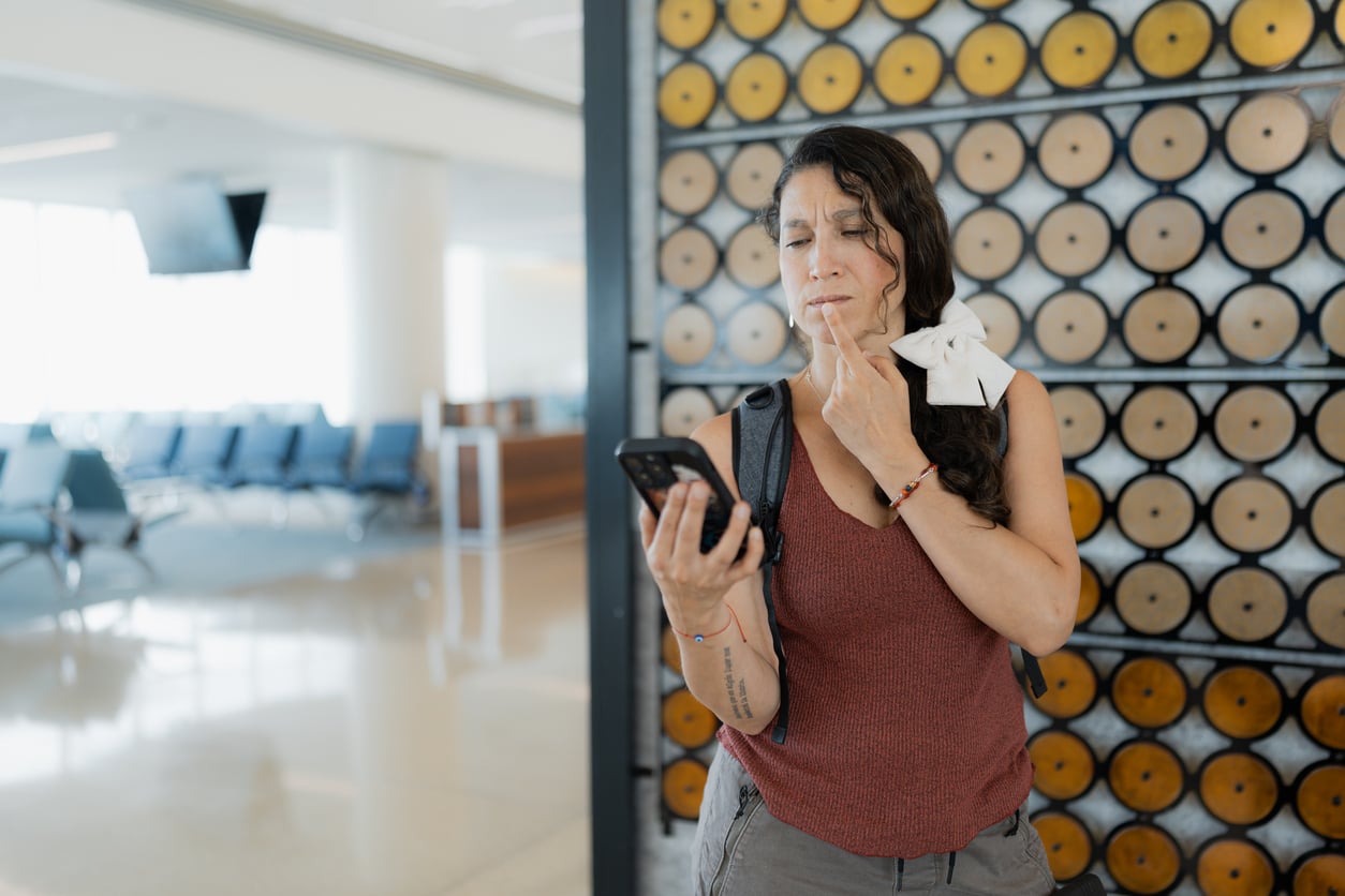 A woman stands in an airport terminal, looking thoughtfully at her phone with a finger on her lips, perhaps checking details about staying at an Airbnb. Empty seats and large windows are visible in the background. She has a white ribbon in her hair.