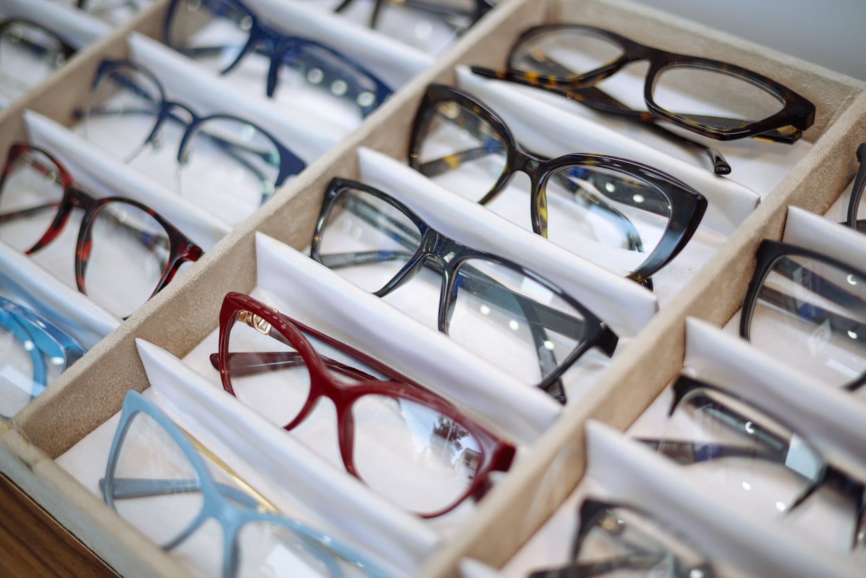 A display case with multiple pairs of eyeglasses in different colors and styles, including red, blue, black, and tortoiseshell frames, arranged neatly in rows.