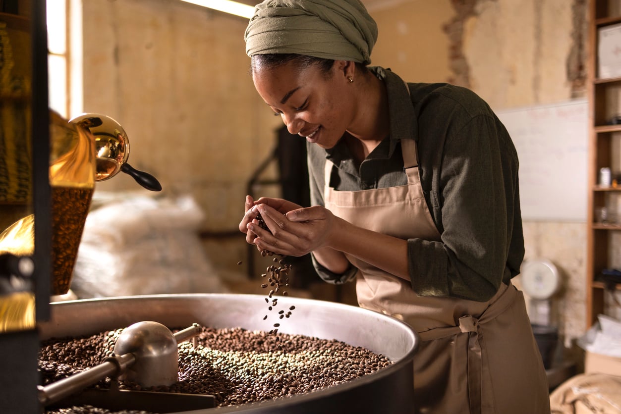 A woman wearing a headscarf and apron smiles as she inspects and smells coffee beans in a large roasting drum inside a rustic workspace.