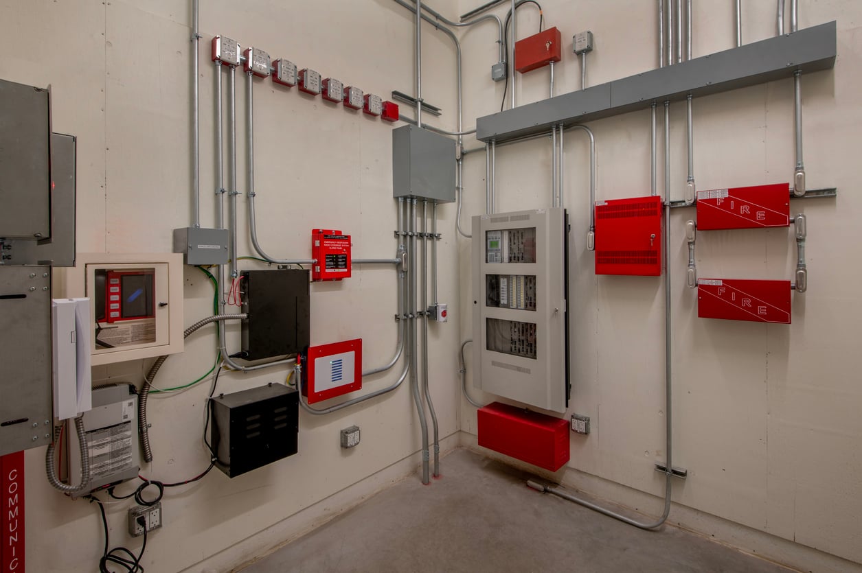 A room with various fire alarm control panels, red emergency equipment, gray electrical conduit pipes, and wiring mounted on light-colored walls. The floor is bare concrete.